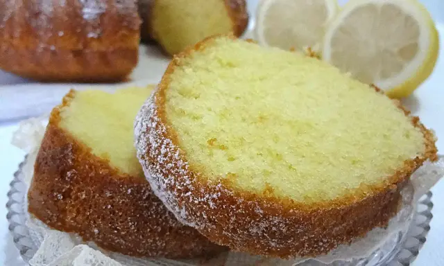 Fette di un soffice ciambellone al limone, spolverate di zucchero a velo, con spicchi di limone sullo sfondo.