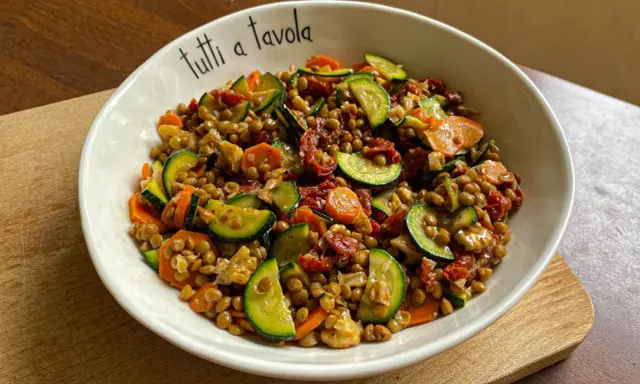 Insalata di lenticchie colorata con zucchine, carote e pomodori secchi, servita in una ciotola con la scritta "tutti a tavola".