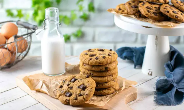 Una pila di biscotti con gocce di cioccolato su un tagliere, accanto a una bottiglia di latte e un cestino di uova.