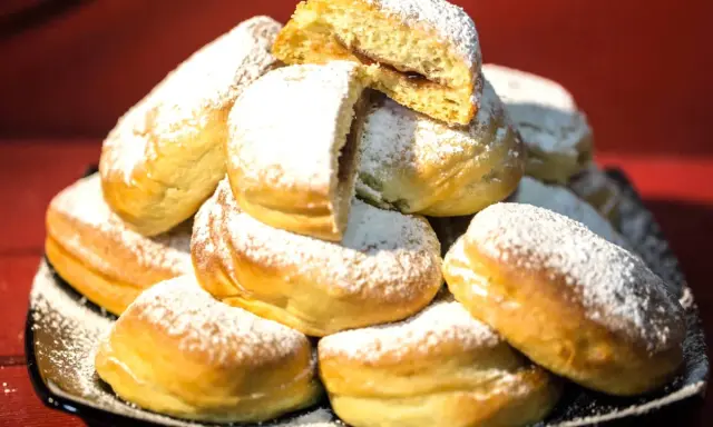 Pile di bomboloni ricetta originale, soffici e cosparsi di zucchero a velo. Uno è tagliato a metà, rivelando un ripieno goloso.