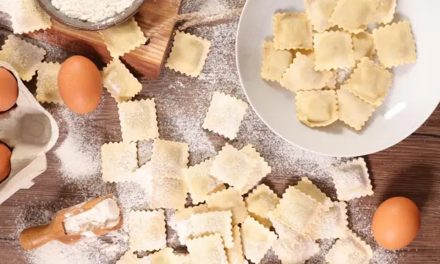 Tanti ravioli fatti in casa sparsi su un tavolo di legno, con farina, uova e una paletta.