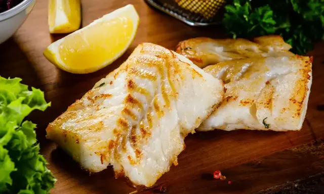 Spigola in padella al vino bianco, dorata e succulenta, servita con limone e insalata fresca su tagliere di legno.