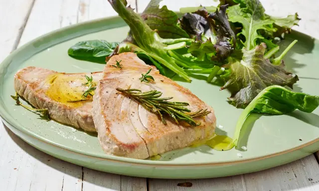 Due fette di tonno in padella, condite con olio e rosmarino, servite con insalata fresca su un piatto verde.