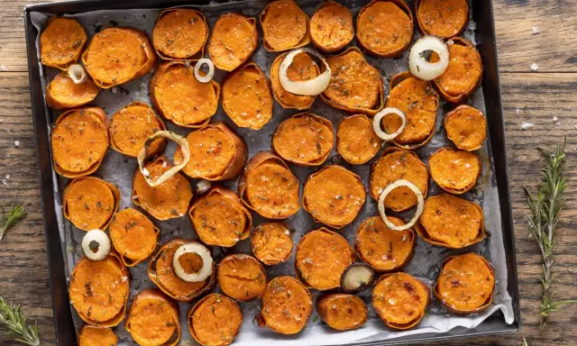 Fette di patate dolci al forno, dorate e condite con erbe aromatiche e sale, disposte su una teglia con anelli di cipolla.