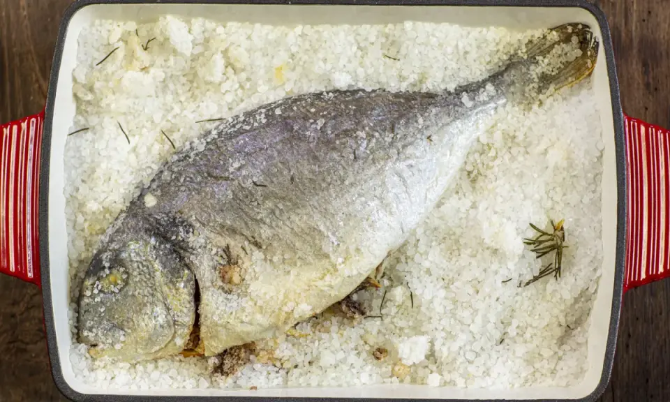 Orata al sale, intera e dorata, adagiata su un letto di sale grosso e rosmarino in una teglia rossa.