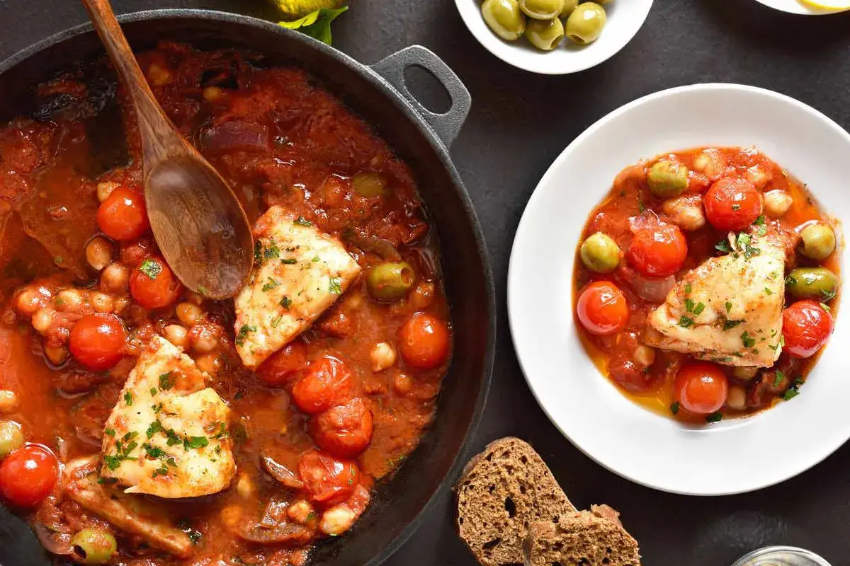 Merluzzo in umido con pomodorini, olive e ceci, servito in una padella e in un piatto, con pane scuro.