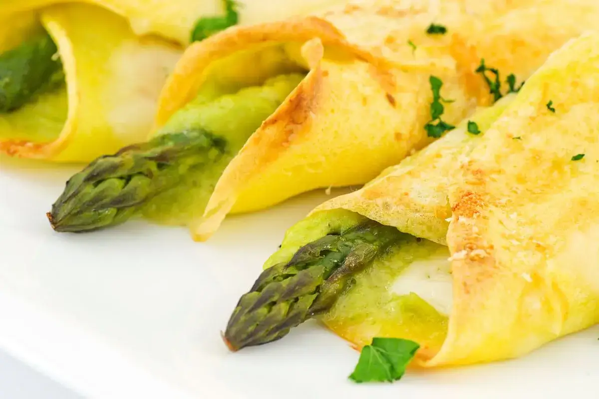 Crespelle con asparagi e formaggio, guarnite con prezzemolo fresco, su un piatto bianco.
