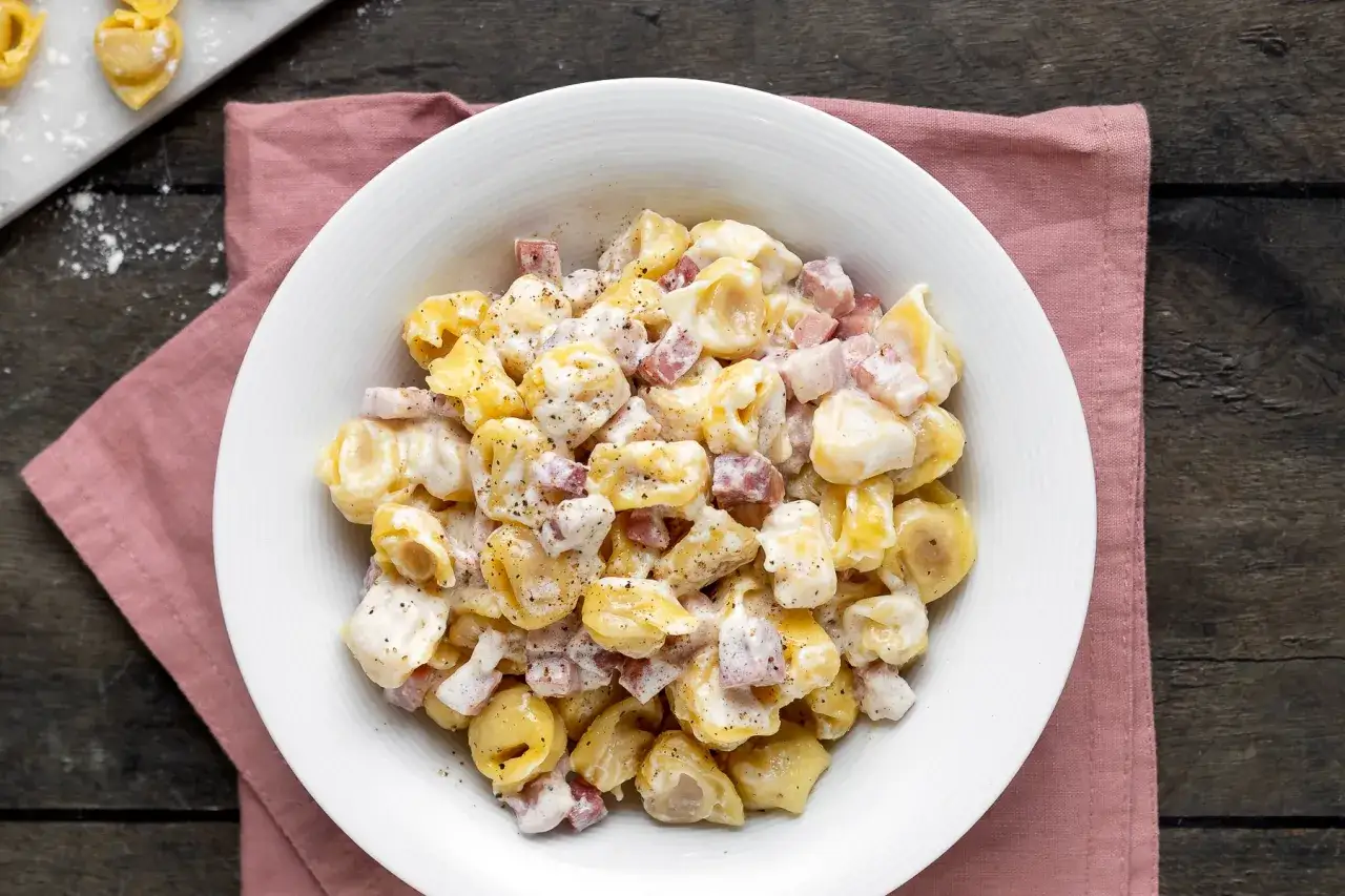 Un piatto invitante di tortellini panna e prosciutto, conditi con pepe nero macinato fresco, su un tovagliolo rosa.