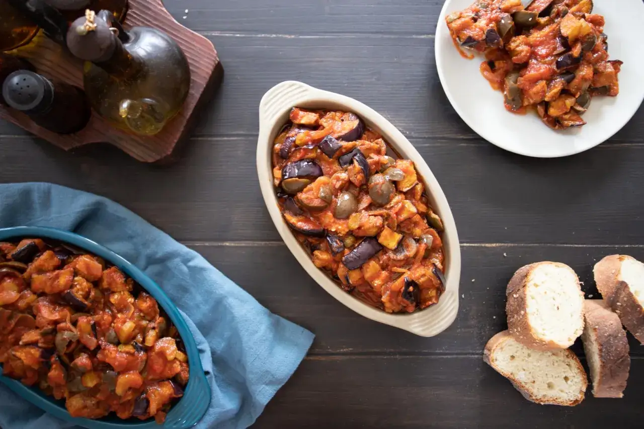 Tre ciotole piene di saporita caponata di melanzane, con pane fresco e condimenti sullo sfondo.