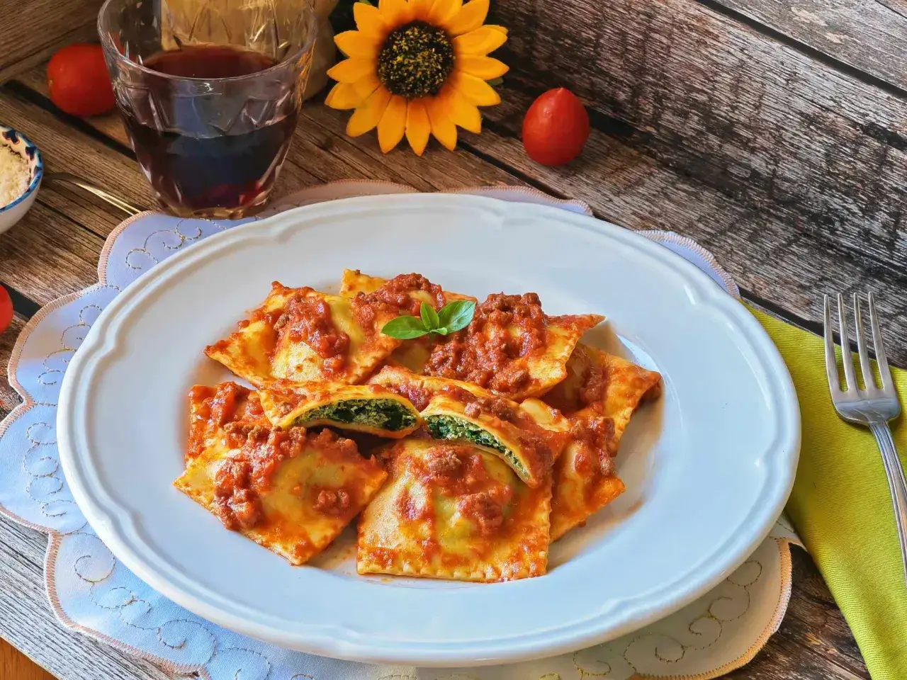 Piatto di ravioli ricotta e spinaci conditi con sugo di carne e basilico, accompagnati da un bicchiere di vino rosso.