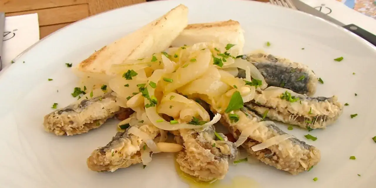 Sarde in saor: pesciolini fritti su un letto di cipolle caramellate, guarniti con prezzemolo e accompagnati da pane tostato.