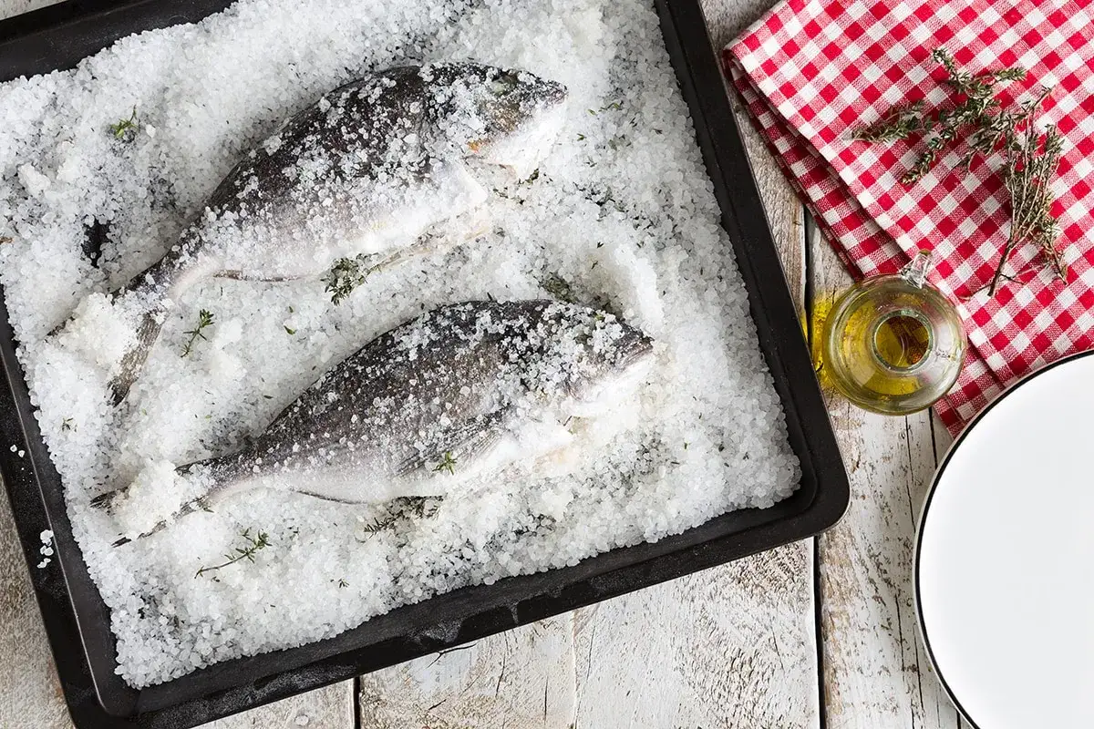 Orata al sale pronta per la cottura, adagiata su un letto di sale grosso con erbe aromatiche. Accanto, un piattino d'olio e un tovagliolo a quadri rossi.