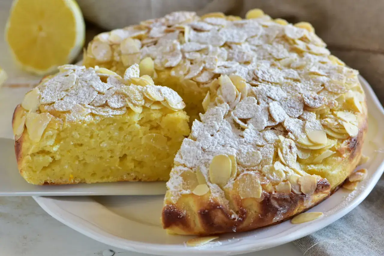 Torta soffice caprese al limone, cosparsa di mandorle a lamelle e zucchero a velo. Un limone intero sullo sfondo.