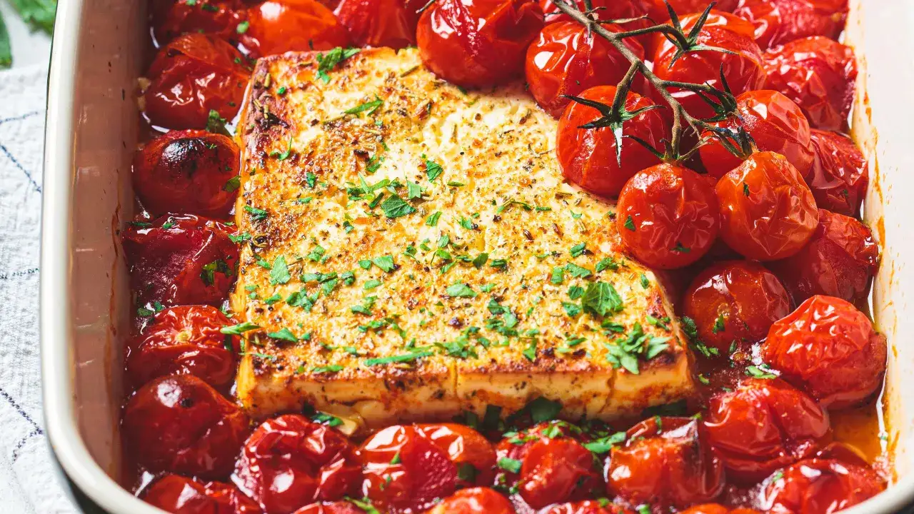 Un blocco dorato di feta al forno, circondato da pomodorini ciliegino succosi e cosparso di erbe fresche.