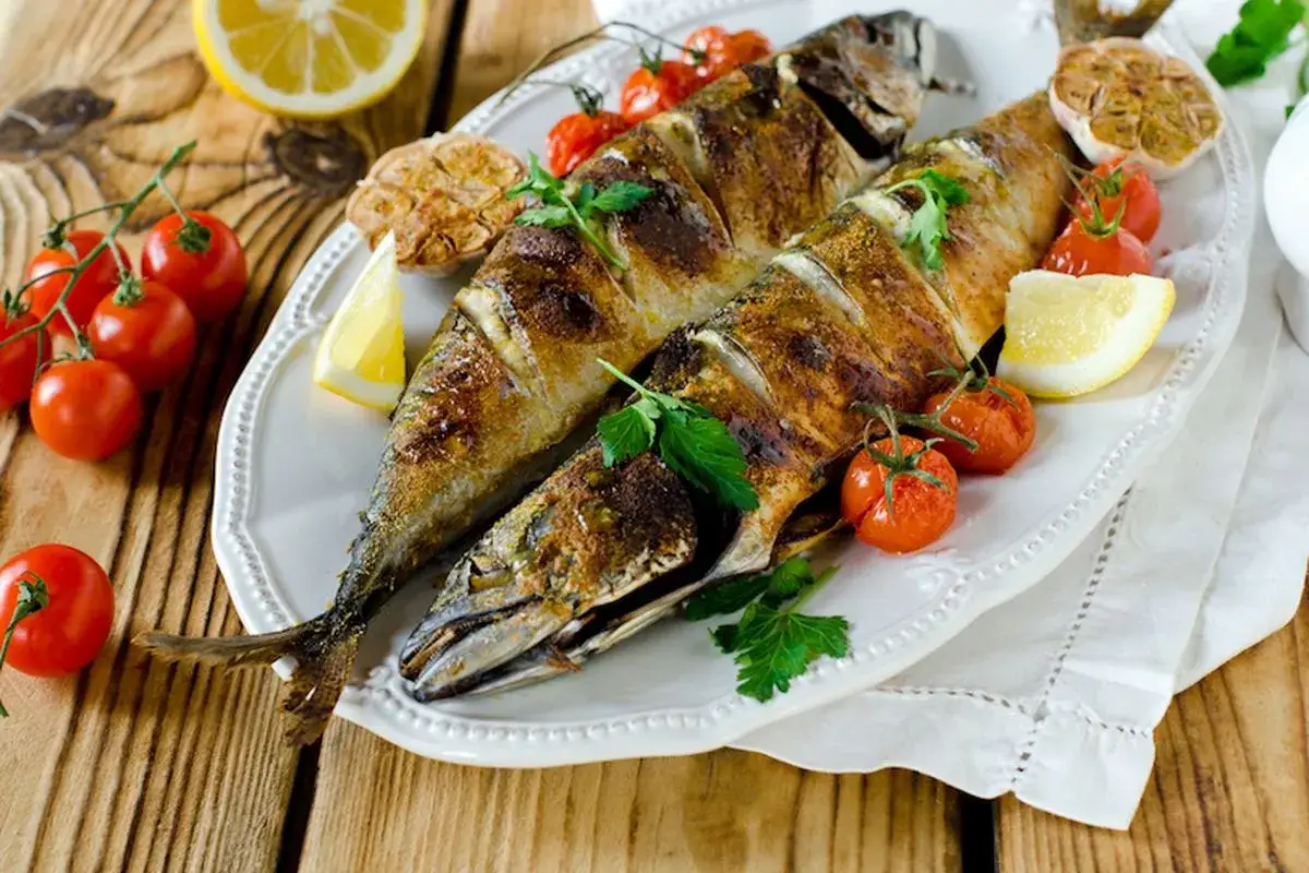 Due filetti di sgombro al forno, dorati e succulenti, serviti con pomodorini, aglio e limone su un piatto bianco.
