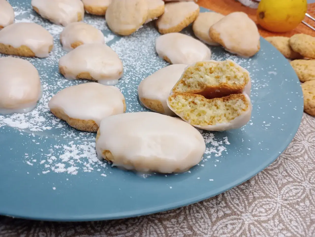 Biscotti al limone glassati, uno spezzato rivela un interno soffice e profumato. Un limone sullo sfondo suggerisce la freschezza.