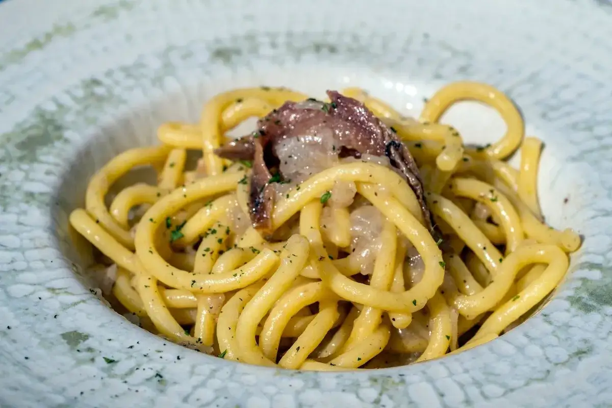 Un piatto di bigoli in salsa, con pasta spessa e condimento a base di acciughe e cipolla, guarnito con prezzemolo tritato.