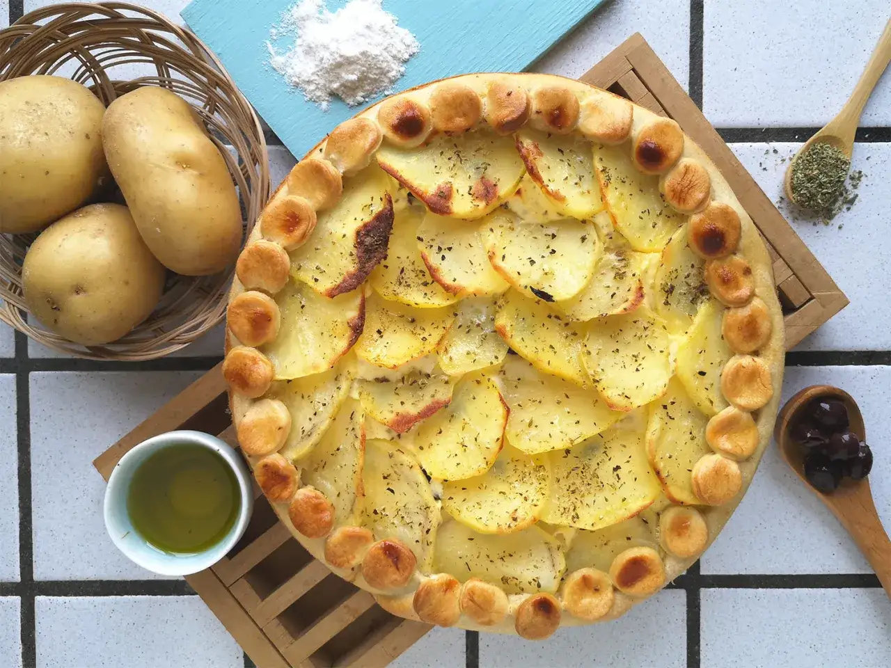 Pizza di patate dorata, con fette sottili di patate e bordi di pasta, accanto a patate fresche, olio e olive.