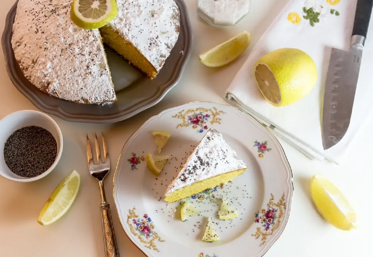 Una fetta di torta al limone velocissima, spolverata di zucchero a velo e semi di papavero, guarnita con spicchi di limone fresco.