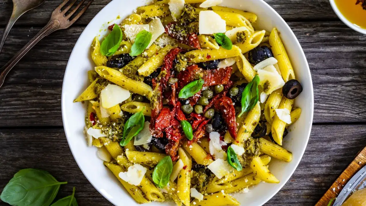 Penne al pesto con pomodori secchi, olive nere, capperi e scaglie di Parmigiano. Un piatto estivo invitante.