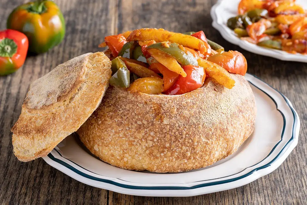 Un pane scavato ripieno di un gustoso stufato di pipi e patate, con una fetta di pane croccante a lato.