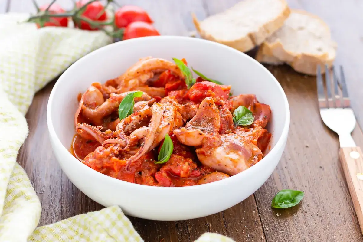 Totani in umido con pomodoro e basilico, serviti in una ciotola bianca, con pane e forchetta sullo sfondo.