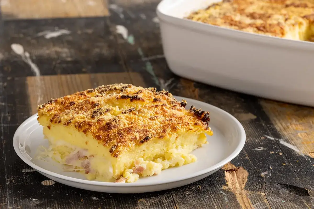 Una fetta di sformato di patate dorato e croccante, con prosciutto e formaggio filante all'interno, servito su un piatto bianco.