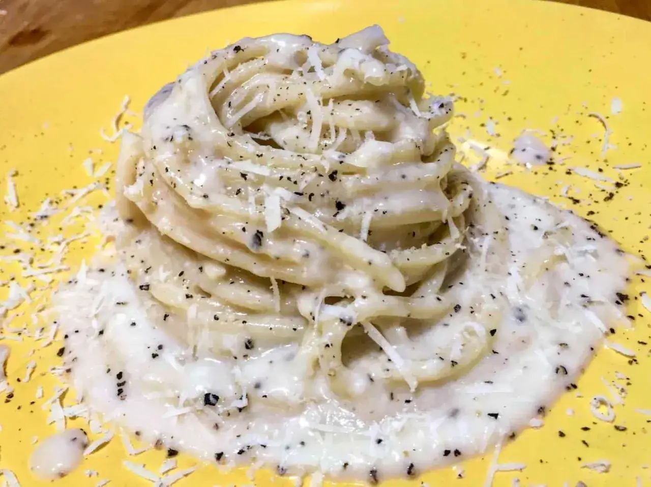 Un piatto di spaghetti al cacio e pepe, cremosi e ricchi di pepe nero macinato, serviti su un piatto giallo brillante.