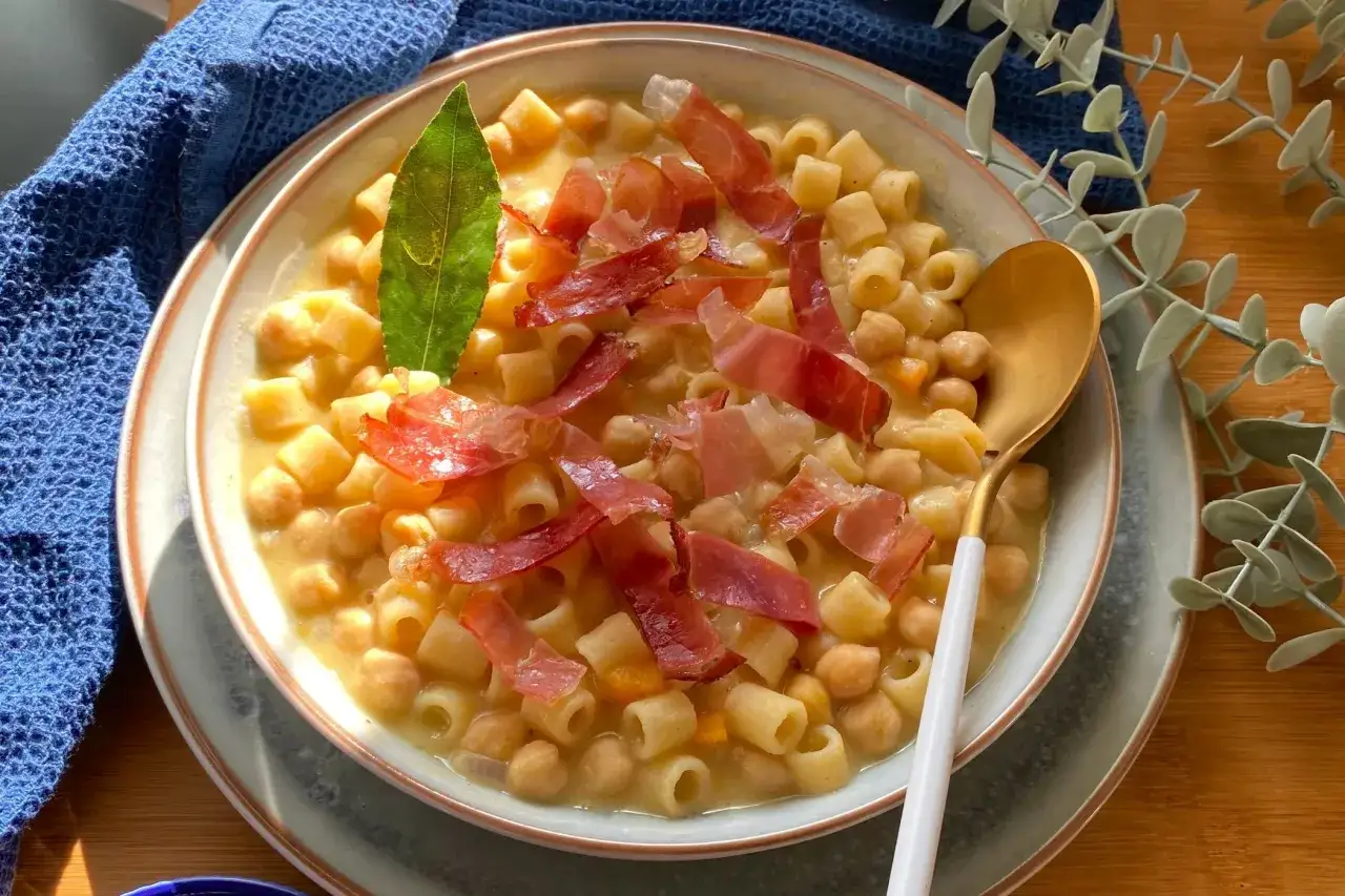 Piatto fumante di pasta e ceci in scatola, guarnito con prosciutto crudo e una foglia di alloro.