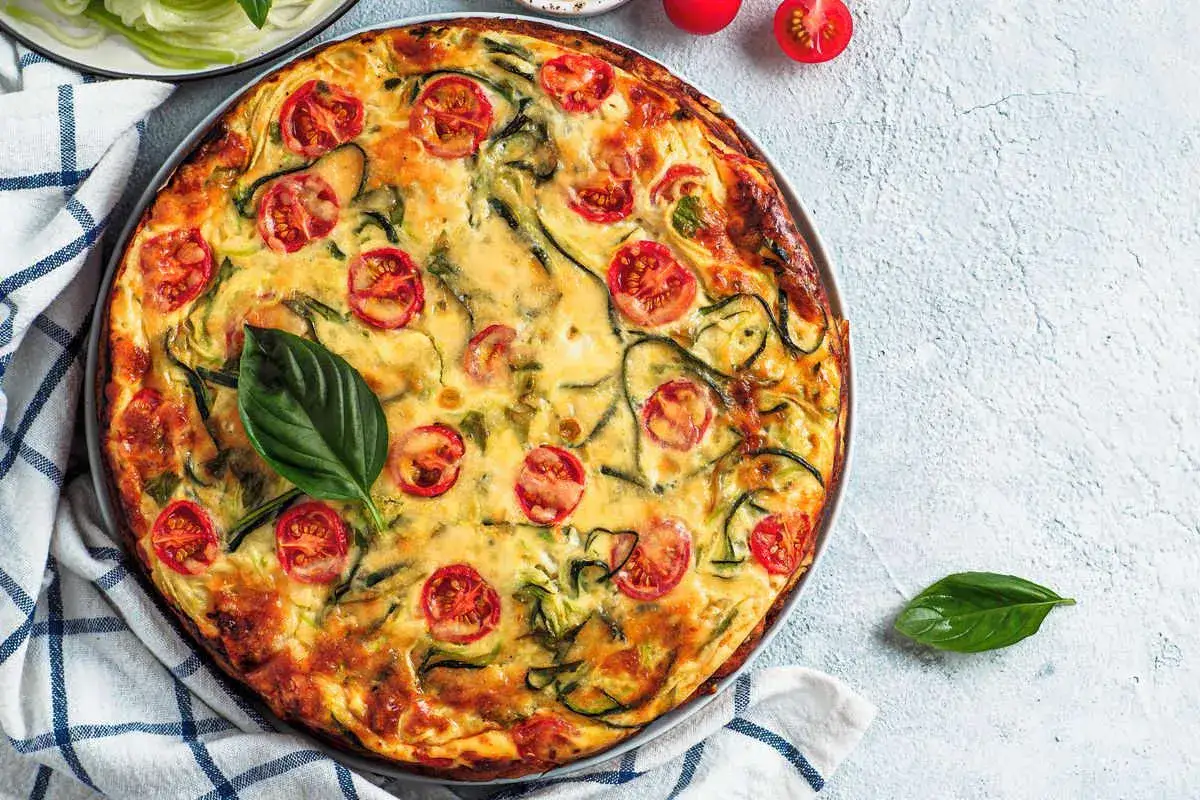 Torta salata vegana dorata con pomodorini e zucchine a spirale, guarnita con basilico fresco.