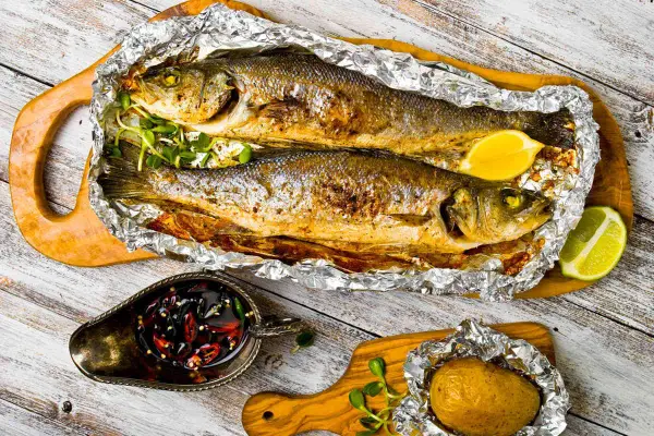 Due branzini cotti al cartoccio, dorati e profumati, serviti con limone, patata al cartoccio e salsa piccante.