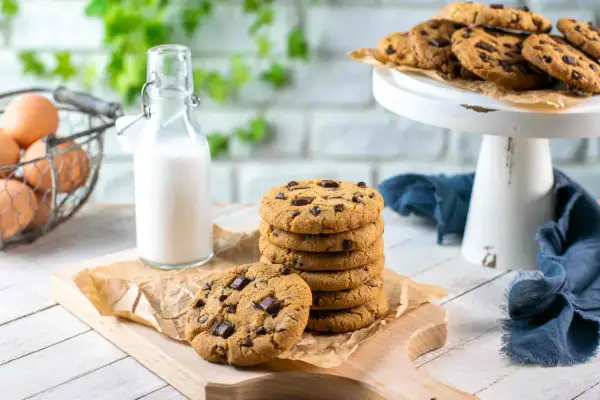 Una pila di biscotti con gocce di cioccolato su un tagliere, accanto a una bottiglia di latte e un cestino di uova.